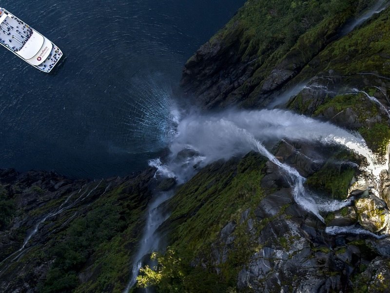 A cruise boat beside a waterfall falling into a fiord, seen from above