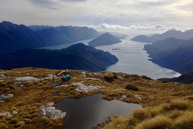 A helicopter parked on a tussock ridge beside a tarn, a long fiord and its arms below